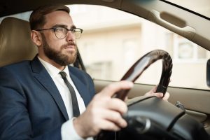 Young successful businessman in eyeglasses and in suit sitting in luxury car and driving